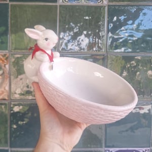 Puede incluir: Un cuenco de cerámica blanca con forma de cesta ovalada y un patrón de tejido texturizado en el exterior. Una figura de conejo de cerámica blanca con mejillas rosadas y una cinta roja fina atada al cuello está unida al lado del cuenco.