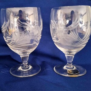 May include: Two clear glass wine glasses with etched floral designs. The glasses have a round bowl and a tall stem with a wide base. The glasses are made by Waterford Crystal.