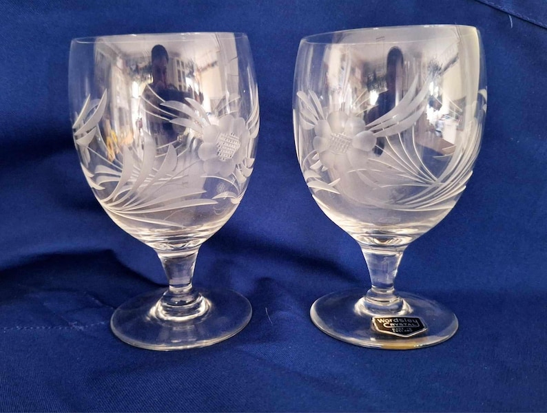 May include: Two clear glass wine glasses with etched floral designs. The glasses have a round bowl and a tall stem with a wide base. The glasses are made by Waterford Crystal.