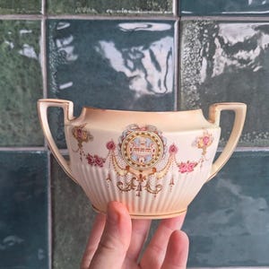 May include: Vintage cream-coloured sugar bowl with two handles and ornate detailing. The bowl has a central medallion with floral and garland accents in pink, blue, and gold. The bowl has a ribbed texture. The bowl is held by a hand.