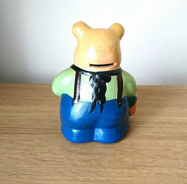 May include: A ceramic piggy bank shaped like a bear wearing blue overalls, a green shirt, and a black bow tie. The bear has a slot on its back for coins.