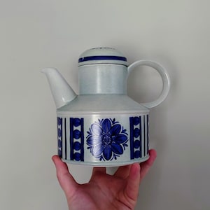 Op de afbeelding: Een blauwe en witte keramische theepot met een bloemenmotief. De theepot heeft een rond lichaam en een tuit aan de zijkant. De handgreep zit bovenop de theepot.