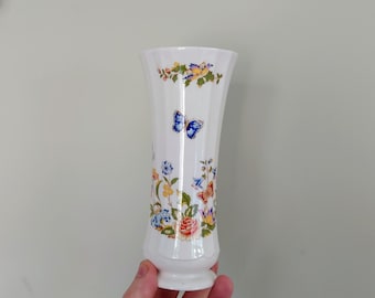 Puede incluir: Jarrón de cerámica blanco con un diseño floral y de mariposa. El jarrón tiene una parte superior acampanada y una base redonda.