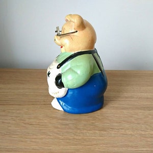 May include: A ceramic piggy bank figurine wearing glasses, a green shirt, and blue overalls. The piggy bank is holding a white object in its hands.
