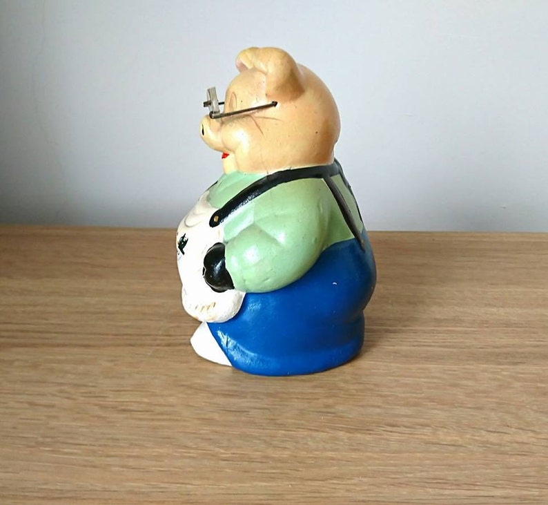 May include: A ceramic piggy bank figurine wearing glasses, a green shirt, and blue overalls. The piggy bank is holding a white object in its hands.
