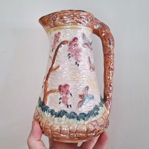 Pode incluir: Um jarro de cerâmica vintage com uma alça texturizada marrom e um corpo branco. O jarro é decorado com um desenho floral e pássaros voando em tons de rosa, azul e verde.