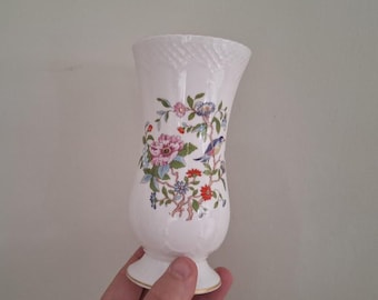 Puede incluir: Jarrón de cerámica blanca con la parte superior acampanada y base de pedestal. El jarrón presenta un diseño floral y de pájaros en rosa, azul y naranja. El borde tiene un patrón texturizado y la base tiene un ribete dorado.