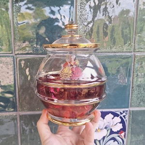 May include: A clear glass jar with a removable lid, featuring a gold-trimmed rim and base. The jar contains a deep red liquid and displays a colorful illustration of two figures in period clothing on the front surface of the glass container.