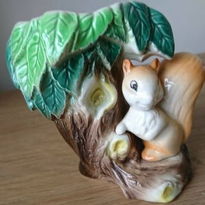 May include: A ceramic squirrel planter with a brown tree trunk and green leaves. The squirrel is white with brown accents and has a bushy tail.