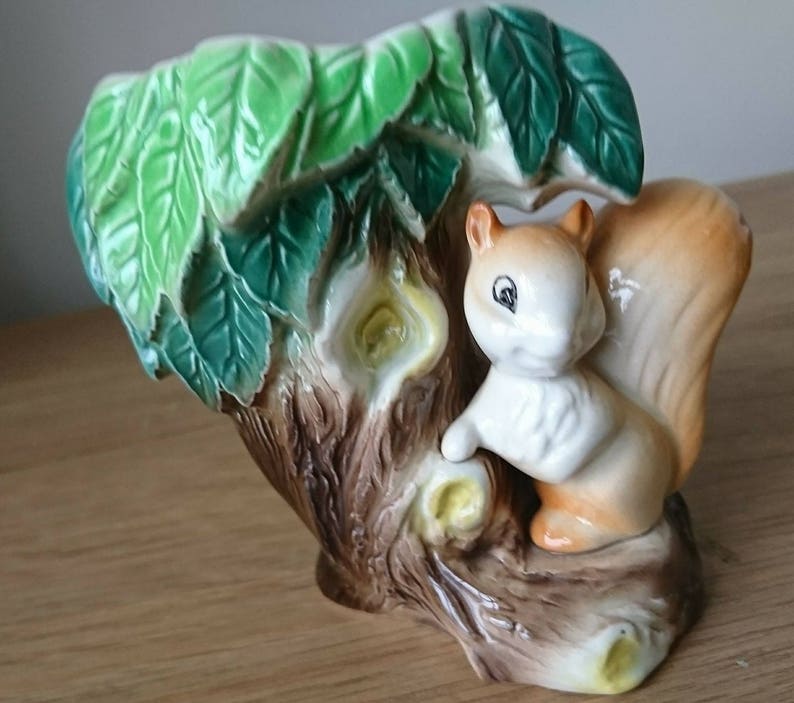 May include: A ceramic squirrel planter with a brown tree trunk and green leaves. The squirrel is white with brown accents and has a bushy tail.