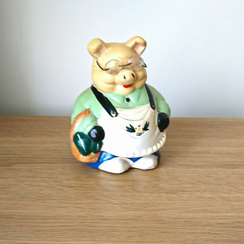 May include: A ceramic piggy bank shaped like a pig wearing a green shirt, an apron, and glasses. The pig is holding a green gardening tool in each hand.