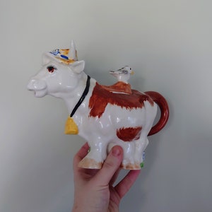 Può includere: Una teiera di ceramica bianca a forma di mucca con macchie marroni, che indossa un cappello giallo e blu e un campanello giallo. Un piccolo uccello bianco è seduto sulla schiena della mucca.
