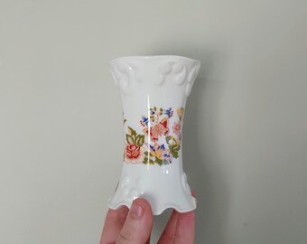 Puede incluir: Un jarrón de porcelana blanca con un borde rizado y un diseño floral con mariposas y flores en tonos rosa, amarillo, azul y verde.