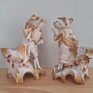 May include: A pair of antique porcelain planters, each featuring a figure in period clothing with a bird perched on their shoulder. The planters are white with gold and brown accents, and floral and leaf details.
