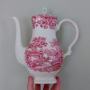 May include: A white ceramic teapot with a pink floral design. The teapot has a round body and a spout on the side. The handle is on the opposite side of the spout.