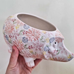 Può includere: Fioriera a forma di riccio in ceramica bianca con un motivo floreale rosa, rosso, giallo e blu. La fioriera ha un'ampia apertura per le piante ed è sostenuta da quattro piedini. Il viso del riccio ha un piccolo occhio nero e un lungo muso.