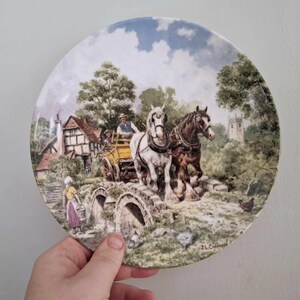 May include: Decorative plate featuring a pastoral scene with a horse-drawn carriage crossing a stone bridge. The plate depicts a village with a church, trees, and figures. The artwork is signed by J.L. Chapman.
