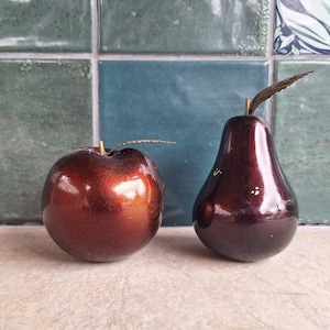 Può includere: Due sculture decorative di frutta: una mela e una pera lucide, marrone-rossastro. La pera è di un colore bordeaux intenso. Entrambe hanno steli e foglie metalliche. Lo sfondo presenta piastrelle verdi e blu.