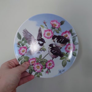Könnte beinhalten: Ein weißer Keramikteller mit blauem Hintergrund und einem Blumendesign mit rosa Rosen und drei schwarz-weißen Vögeln. Der Teller ist signiert mit "Jeff West".