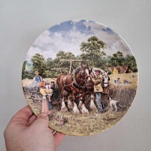 May include: Decorative plate featuring a pastoral scene with two brown horses pulling a cart. A farmer and a dog are also present. The plate has a blue sky and green trees in the background. The plate is held by a hand.