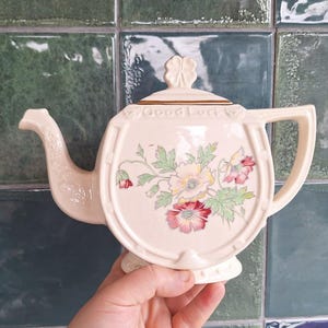 May include: Cream-colored ceramic teapot with a horseshoe shape and floral design. The teapot features a lid with a clover, a spout, and a handle. The words "Good Luck" are written on the rim. The floral design includes red, pink, and yellow flowers.