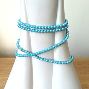 Pode incluir: Um fio de contas de pérola azul claro iridescentes enroladas em torno de um vaso branco. As contas são pequenas e redondas, e têm um brilho sutil.