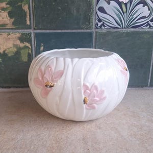 Peut inclure: Un bol en céramique blanche de forme arrondie et côtelée, orné de motifs de fleurs de lotus roses en relief en trois dimensions avec des centres dorés. Le bol présente une finition lisse et brillante ainsi qu'une large ouverture supérieure.