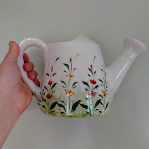 Puede incluir: Regadera de cerámica blanca con un diseño floral con hojas verdes, flores rojas, amarillas y naranjas.