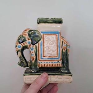 Op de afbeelding: Een keramieken olifantenbeeldje in groen, blauw en beige. De olifant heeft gedetailleerde kenmerken en decoratieve patronen. Het beeldje wordt door een hand vastgehouden en lijkt een decoratief item te zijn.