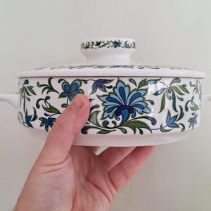 Op de afbeelding: Witte keramische ovenschaal met een blauw en groen bloemenpatroon. De schaal heeft een deksel met een knop.