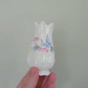 Può includere: Un piccolo vaso in ceramica bianca con un motivo floreale. Il vaso ha una superficie testurizzata e un delicato disegno di farfalla con accenti rosa, blu e verdi.