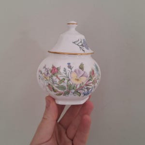May include: A white porcelain sugar bowl with a lid, featuring a colourful botanical pattern of pansies, roses, and thistles. The rim of the lid and the base of the bowl are accented with a thin gold trim, and the lid has a small, rounded finial.