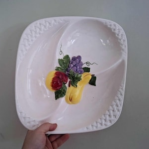 May include: White ceramic serving dish with a woven design around the edge. The dish is divided into three sections and features a painted design of fruit, including a pear, grapes, and a yellow squash.