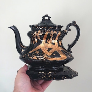 May include: Black teapot with gold floral design and a decorative base. The teapot has a spout and a handle.