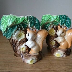 May include: Two ceramic squirrel planter vases with green leaves and brown tree trunks. Each vase features a squirrel with a bushy tail and brown and white fur.