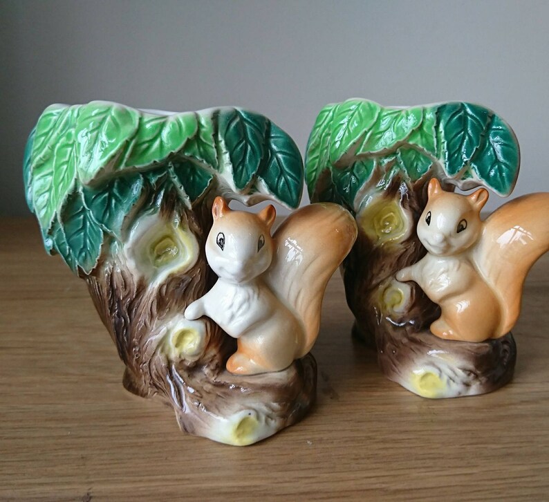 May include: Two ceramic squirrel planter vases with green leaves and brown tree trunks. Each vase features a squirrel with a bushy tail and brown and white fur.