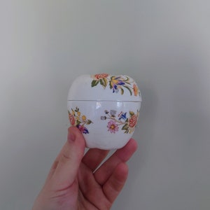 May include: A white ceramic trinket box shaped like an apple with a floral design. The flowers are pink, yellow, blue, and red.