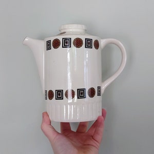 May include: Cream-colored ceramic coffee pot with a lid and handle. The pot is decorated with a repeating pattern of brown and black floral designs. The pot is held in a hand.