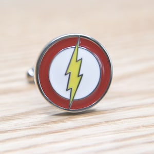 May include: A pair of cufflinks featuring the Flash logo. The logo is a yellow lightning bolt inside a red circle with a white background.
