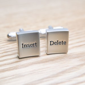 May include: Silver-coloured cufflinks featuring the words "Insert" and "Delete" on square faces. The cufflinks are presented on a light-coloured wooden surface, showcasing a modern design.