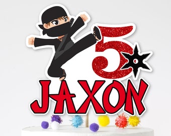 May include: A white cake with a red and black ninja cake topper. The topper features a cartoon ninja kicking a red number 5 with a black star. The cake topper also includes the name Jaxon in red letters.
