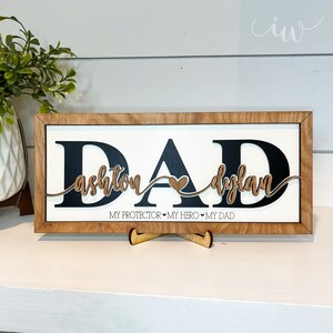 May include: Wooden sign with the word "DAD" in black letters with the names "Ashton" and "Dylan" in brown letters on a white background. The sign has a brown wood frame and reads "My Protector, My Hero, My Dad".