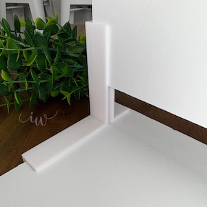 May include: A white, right-angled display stand, possibly for product photography. The stand is made of a solid, opaque material and is positioned on a white surface. Green foliage is visible in the background, along with the cursive text "ciw".