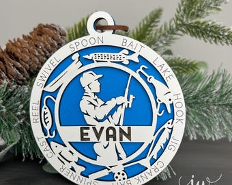 May include: A round, layered ornament with a blue interior and white exterior. The design features a fisherman with the name "EVAN" and fishing-related words like "spoon" and "hook." A brown leather loop is at the top for hanging. The ornament is decorated with pine needles.