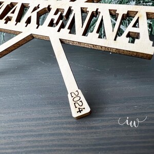 May include: A wooden ornament with the name "AKENNA" cut out in the centre. The ornament is shaped like a star with a pointed bottom that reads "2024".