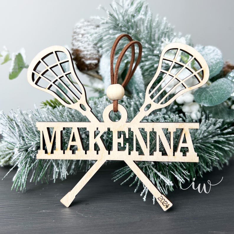 May include: Wooden Christmas bauble featuring two lacrosse sticks crossed over a banner with the name "MAKENNA" and the year "2024" inscribed on the bottom.
