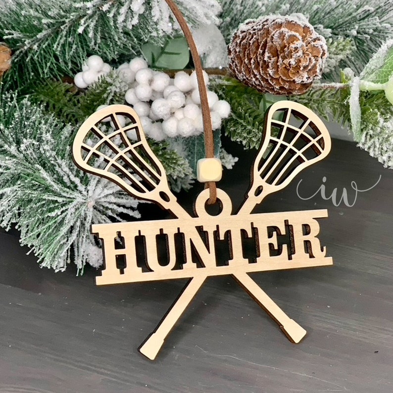 May include: Wooden Christmas bauble featuring two lacrosse sticks crossed with the name "Hunter" in a banner below. The bauble is brown and has a white string for hanging.