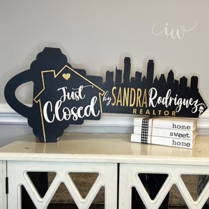 May include: Black and gold key-shaped sign with the text "Just Closed" and "by SANDRA Rodriguez REALTOR". The sign is propped up on a white table with a stack of books that say "home sweet home".