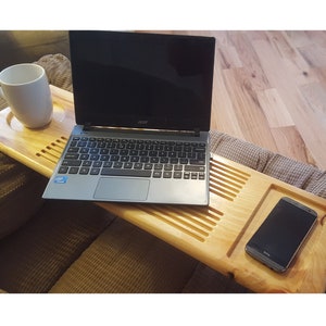 May include: A light-colored wooden laptop tray with a built-in cup holder and a slot for a phone. An Acer laptop and a white mug are on the tray. The tray is resting on a brown couch.
