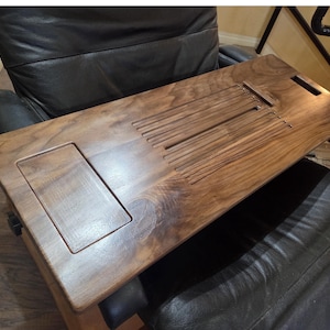 May include: A rectangular dark brown wooden lap desk featuring a smooth surface with a recessed rectangular area on the left side and a series of parallel horizontal grooves in the center, designed to hold a tablet or phone upright.